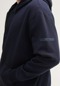 Navyblauwe hoodie van zacht materiaal, met een rits aan de voorkant, kangoeroezakken en een logo-opdruk op de linkermouw.