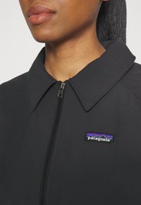 Černá bunda se zipovým uzavíráním a velkým límcem. Na hrudi je barevná nášivka Patagonia. Hladká, texturovaná látka.