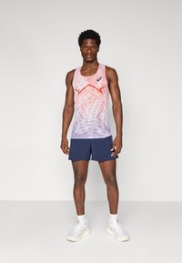 Mouwloze sporttanktop in een gradiënt van roze en paars gaas, met geometrische patronen. Gecombineerd met marineblauwe shorts en witte hardloopschoenen.