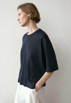 Jeune personne aux cheveux courts portant un T-shirt noir ample et un pantalon blanc, debout de profil avec une main dans la poche, contre un mur uni.