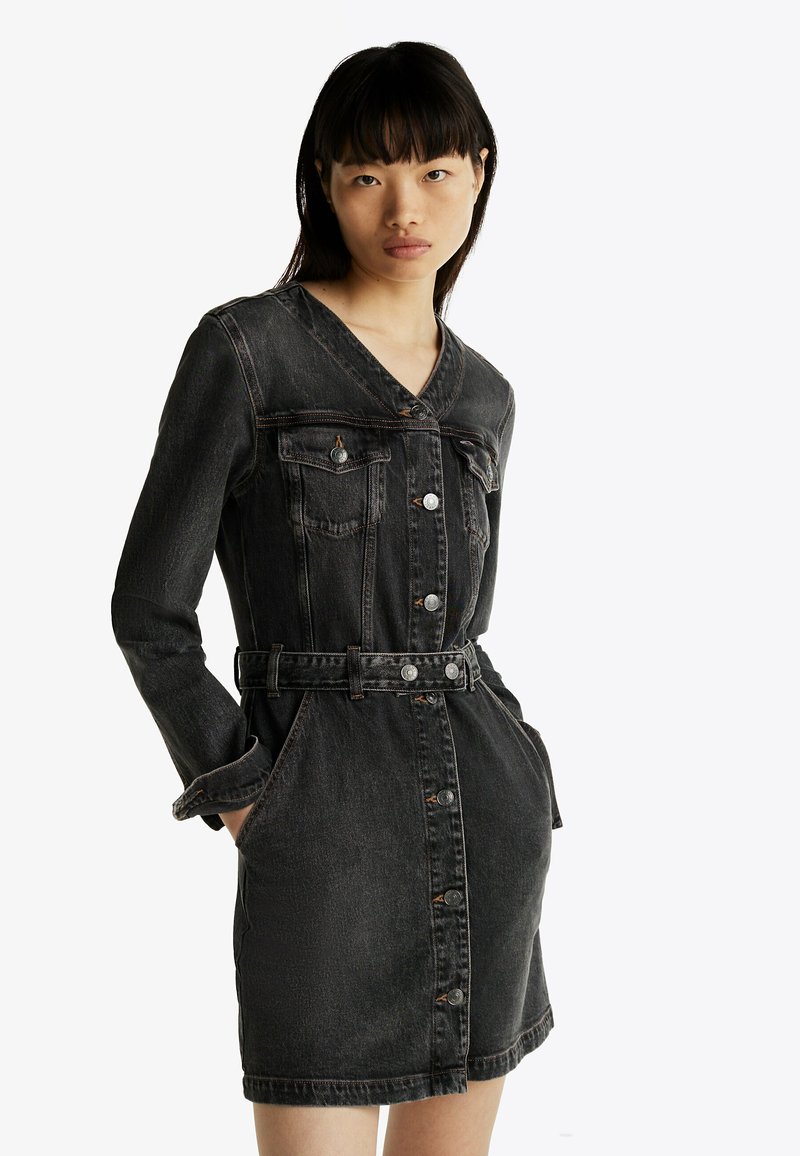 Abito in denim nero con maniche lunghe, scollatura a V, bottoni frontali, tasche sul petto e vita con cintura. Presenta una texture leggermente strappata.