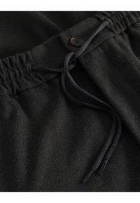 Pantalons de survêtement noirs en tissu texturé, avec une taille élastique dotée d'un cordon de serrage et un détail de bouton marron.
