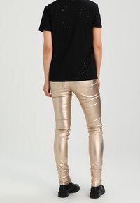 Camiseta negra con detalles desgastados combinada con pantalones ajustados metálicos dorados y botines negros, vista desde atrás.