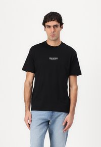 Sort bomulds T-shirt med korte ærmer; har et hvidt trykt tekstlogo på brystet. Model iført lysblå denim jeans.