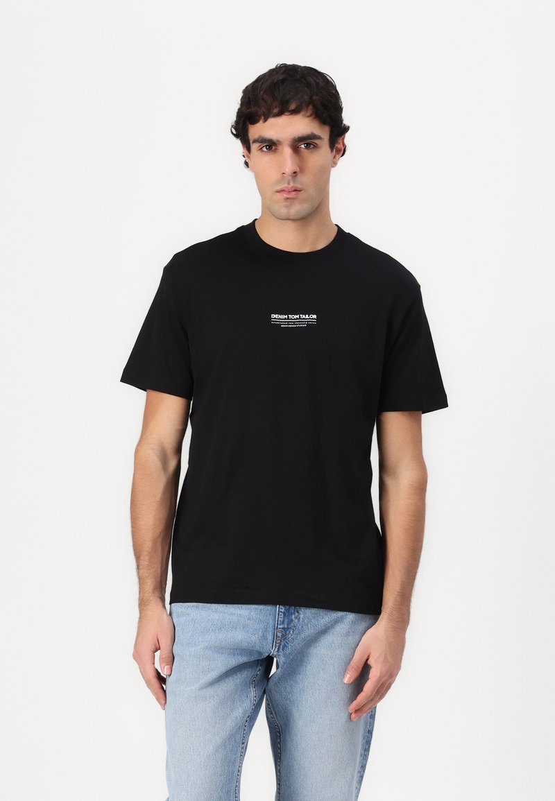 Sort bomulds T-shirt med korte ærmer; har et hvidt trykt tekstlogo på brystet. Model iført lysblå denim jeans.
