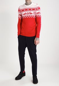 Uomo che indossa un maglione di Natale a righe rosse e bianche, pantaloni neri, scarpe nere, in piedi con le mani nelle tasche su uno sfondo semplice.