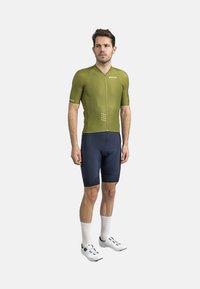 Maillot de cyclisme à manches courtes vert olive avec une fermeture éclair à l'avant, associé à un short de cyclisme bleu marine et des chaussettes blanches. Présente un branding subtil.