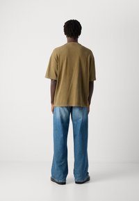 Mand med korte dreadlocks iført en oversized olivengrøn t-shirt, løse blå jeans og sorte sko, vendt mod en ensfarvet hvid væg.