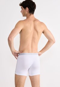 Boxer in cotone bianco con cintura grigia, design aderente. Modello mostrato di spalle, che evidenzia una texture liscia e uno stile minimalista.