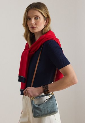 Γυναίκα που φοράει μπλε κοντομάνικη μπλούζα με κόκκινο πουλόβερ περασμένο, κρατάει μικρή τσάντα από τζιν, κρεμ παντελόνι και βραχιόλι από κέλυφος χελώνας.