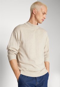 Jersey de cuello alto beige hecho de un suave tejido de punto, con un cuello ajustado y puños acanalados. Se lleva con vaqueros azul oscuro.