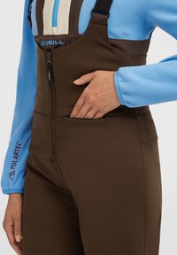 Salopette marrone con tasca frontale con zip, realizzata in materiale liscio. Sotto è indossata una giacca in pile blu con zip e logo.