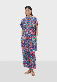 Un mono de estampado floral colorido con cuello de camisa, mangas cortas, bolsillos y un corte relajado, fabricado en una tela suave y ligera.