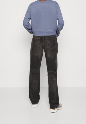 Personne portant un jean noir délavé à jambes droites, un sweat-shirt bleu à manches longues et des baskets blanches, debout, les mains sur les hanches, de dos.