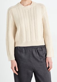 Cable-knit cream sweater featuring long sleeves, ribbed cuffs, and hem; cropped design with textured detailing. Worn over dark gray trousers.