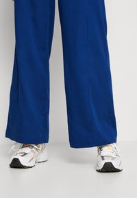 Oříznutý pohled na široké modré kalhoty z hladké tkaniny, kombinované s bílými teniskami s šedými a výraznými detaily.