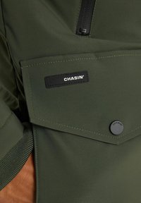 Giacca verde oliva con una tasca con cerniera, una tasca con patta con un'etichetta nera con la scritta "CHASIN'" e dettagli testurizzati sull'orlo.