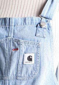 Lichtblauwe denimsalopetten met een wit label met een zwart logo, metalen sluitingen en een voorzak. Geweven stof met subtiele stiksels.