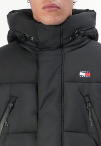 Uomo che indossa un giubbotto imbottito nero con collo alto, tasche con zip e logo Tommy Jeans sul petto.