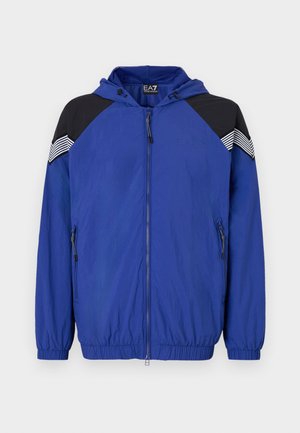Chaqueta ligera azul con capucha, con acentos en negro en los hombros y detalles a rayas. Frente con cremallera y puños elásticos. Textura suave.