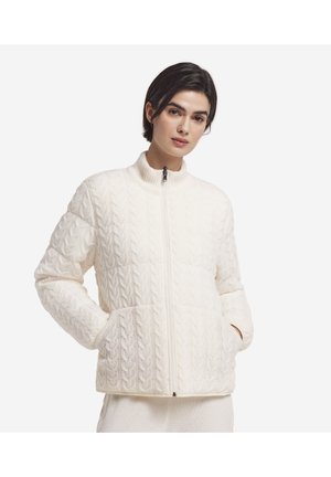 Kvinde iført en hvid kabelstrikket trøje med lynlås og lommer, stående mod en ensfarvet hvid baggrund.