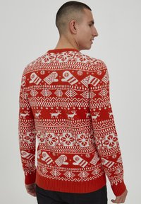 Pull rouge en maille avec des motifs de Noël blancs, incluant des rennes, des flocons de neige et des mitaines. Col rond et poignets côtelés.