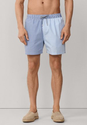 Hombre con bañador corto hasta media pierna con rayas verticales azules y blancas, combinado con alpargatas beige sin cordones.