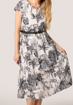 Vrouw draagt een witte midi-jurk met zwarte tropische en dierenprint, korte mouwen, een keyhole-halslijn en een smalle zwarte riem in de taille.