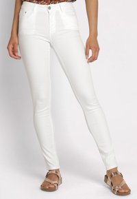 Witte, slim-fit jeans met een gladde textuur, vijf zakken en een middelhoge taille. Gecombineerd met tan gekruiste sandalen.