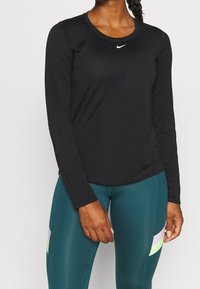 Kvinna som bär en svart långärmad Nike-tröja och tealleggings med pastellrandiga detaljer, stående med händerna avslappnade vid sidorna mot en enfärgad bakgrund.