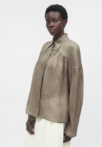 Γυναίκα με κοντά σγουρά μαλλιά φορά χαλαρό, διαφανές μπλουζάκι ταμπ με μαζεμένους ώμους και φαρδιά μανίκια πάνω από λευκή φούστα, ποζάροντας σε λευκό φόντο.