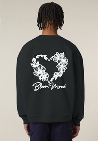 Felpa nera con un design a forma di cuore floreale bianco e il testo "Bloom Mood" sotto. Tessuto morbido, vestibilità comoda, polsini e orlo a coste.