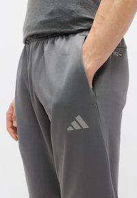 Pelēkas sporta bikses ar elastīgu jostasvietu, sānu kabatām un atstarojošu Adidas logotipu kreisajā kājā. Gluda, viegla auduma.