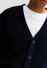 Marinblå stickad kofta med V-hals, med texturerad tyg och två blå knappar framtill, bärs över en vit t-shirt med rund hals.