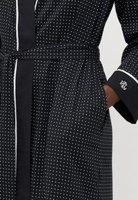 Svart badekåpe med hvite prikker, belted i livet. Har sidelommer og hvite pipinger på ermene med broderte logo-detaling.