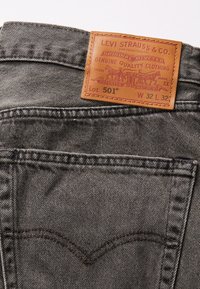 Levi's 501 farkut tummanharmaasta denimmateriaalista, nahkajäljitelmällä varustettu logo ja ompeluakkentteja, sekä erottuva takataskun muotoilu.