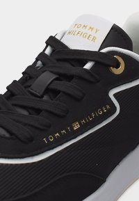 Μαύρο αθλητικό παπούτσι Tommy Hilfiger με μαύρα κορδόνια, χρυσά οπές για τα κορδόνια και χρυσό λογότυπο στο πλάι και στη γλώσσα, λευκή σόλα και επένδυση.