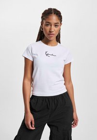 T-shirt bianco in cotone con maniche corte, caratterizzato da un logo nero scritto a mano sul lato sinistro del petto, abbinato a pantaloni cargo neri larghi.