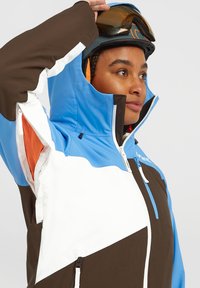 Blauwe, bruine en witte skijas met een hoge kraag, ritssluitingen en een mesh voering. Bevat een capuchon en een kleurgebroken ontwerp.