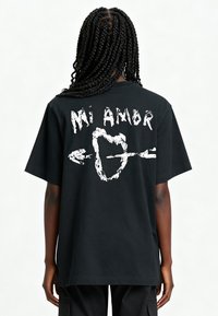 T-shirt en coton noir avec le texte graphique blanc "Mi Amor" et un design de flèche au dos. Coupe oversize avec manches courtes.