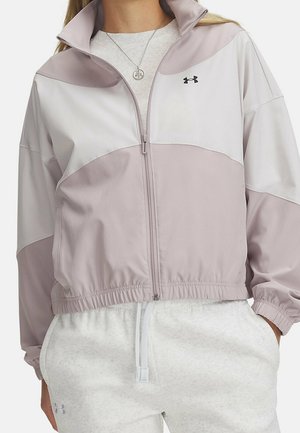 Femme portant une veste zippée violet clair et blanc avec ourlet élastique et logo Under Armour, associée à un pantalon de survêtement gris clair avec cordon de serrage.