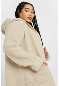 Beige oversized fleecemantel med huva, med en frontficka, strukturerad tyg och avslappnad silhuett. Mjuk och varm design.