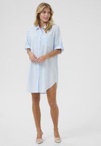 Robe chemise bleu clair en tissu texturé, dotée d'une poche poitrine et de manches retroussables, avec un ourlet légèrement arrondi.