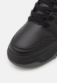 Sort læder sneakers med perforeret tekstur, flade sorte snørebånd og en tykk sort gummisål med et struktureret grebmønster.