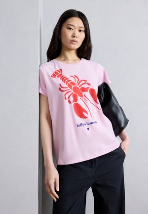 Νεαρή γυναίκα που φοράει ανοιχτό ροζ μπλουζάκι με μεγάλο κόκκινο σχέδιο αστακού και μπλε κείμενο «lollys laundry», κρατάει μαύρη τσάντα και ποζάρει μπροστά από έναν τοίχο.