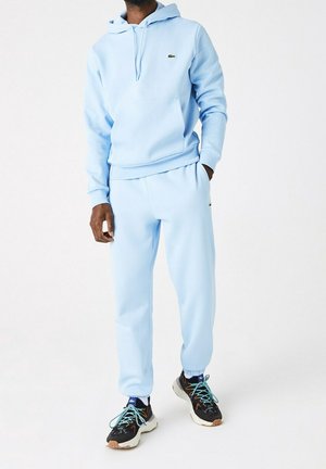 Homme portant un sweat à capuche bleu clair et un pantalon de survêtement assorti avec des baskets noires et blanches, mains dans les poches, debout devant un fond blanc.