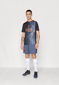 Sportströja i svart och blått med orangea accenter, tillsammans med blå shorts. Funktioner inkluderar sponsorslogotyper och en stilren design.