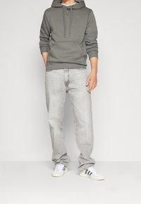 Felpa grigia con tasca frontale, abbinata a jeans grigi chiari. Calzature bianche con strisce nere. Tessuto liscio, vestibilità casual.
