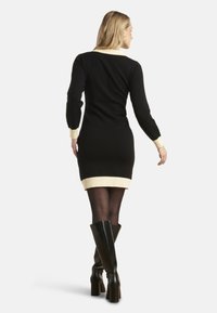 Robe en tricot noire avec des manches longues, accents crème au col et à l'ourlet, silhouette ajustée, associée à des bottes noires montant jusqu'aux genoux.