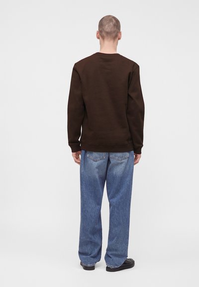 Homme portant une chemise marron foncé à manches longues, un jean bleu ample et des chaussures noires, debout dos à la caméra.